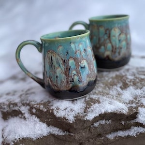 May include: Two ceramic mugs with teal and brown glaze. The mugs have a black base and a curved handle. The mugs are sitting on a rock covered in snow. The mugs are handmade and have a unique design.
