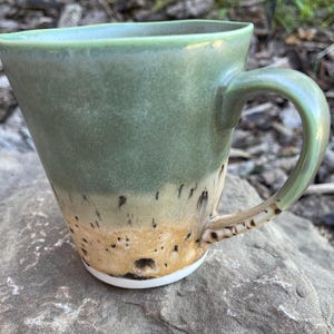 Peut inclure: Une tasse en céramique avec un bord large et évasé ainsi qu'une anse incurvée. La partie supérieure présente un émail vert sauge mat et lisse, tandis que la section inférieure passe à une finition tachetée beige et brune avec une base blanche.