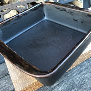 May include: A rectangular, dark grey baking dish with a glossy finish and a brown rim. The dish is empty and appears to be made of ceramic or a similar material. The baking dish is sitting on a wooden surface.