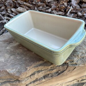 May include: A light green ceramic loaf pan with a white interior. The pan has a slightly textured finish and rounded edges.