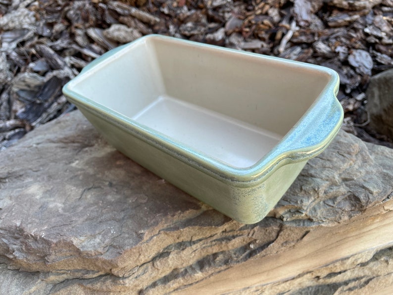 May include: A light green ceramic loaf pan with a white interior. The pan has a slightly textured finish and rounded edges.