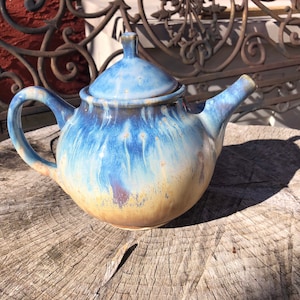 May include: A blue and yellow ceramic teapot with a lid. The teapot has a round body and a curved spout. The teapot is sitting on a wooden surface.