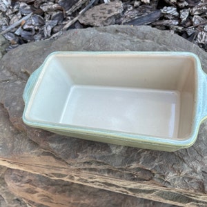 May include: A green ceramic loaf pan with a light beige interior. The pan has two handles on the sides.