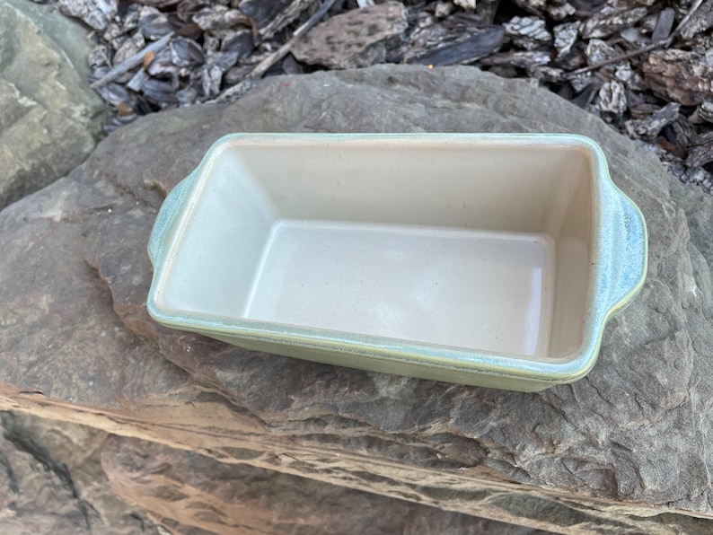 May include: A green ceramic loaf pan with a light beige interior. The pan has two handles on the sides.