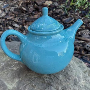 May include: A glossy turquoise ceramic teapot with a rounded body, a curved handle, a pointed spout, and a matching lid with a small knob handle. The teapot features a smooth, reflective surface and a uniform bright blue-green colour throughout.