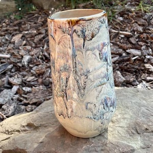 Peut inclure: Un vase en céramique de grande taille avec un motif de glaçure unique. Le vase présente une base crème avec des stries de marron, de bleu et de violet. Le vase cylindrique est exposé sur une surface en pierre naturelle.