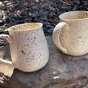 May include: Two speckled ceramic mugs with large handles. The mugs are off-white with brown speckles and sit on a grey stone surface. The mugs are handmade and have a rustic, artisanal appearance.