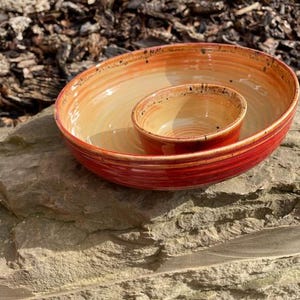 May include: A ceramic serving bowl set, handmade with a red exterior and yellow interior. The smaller bowl sits inside the larger one. The bowls are on a stone surface. The background is blurred, showing wood chips.