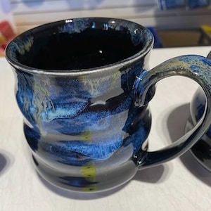 Peut inclure: Une tasse en céramique avec une finition brillante présentant une base noir profond mélangée à des motifs tourbillonnants de bleu cobalt, de bleu clair et de légères touches de jaune. La tasse a un corps arrondi légèrement ondulé.