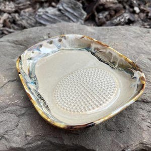 May include: A handmade ceramic garlic grater dish. The oval-shaped dish features a textured, off-white interior and a raised, circular grating surface. The rim has a decorative, multi-coloured glaze with brown, blue, and gold tones.