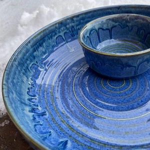 May include: A blue ceramic serving set comprising a large, shallow dish and a smaller bowl. The set has a glossy finish with a textured, layered design. The bowl sits atop the larger dish, creating a cohesive presentation.