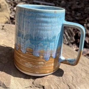 Peut inclure: Une tasse en céramique faite à la main avec une anse rectangulaire. La tasse présente un glaçage bleu et marron, le bleu dégoulinant sur la base marron. La tasse est posée sur une roche.