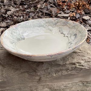 May include: A handmade ceramic bowl with a wide, shallow design. The bowl has a creamy white interior and a rim with a marbled effect of grey and white. The rim has a subtle pinkish-brown edge. The bowl is resting on a stone surface.