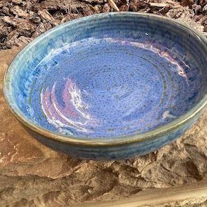 May include: A round ceramic bowl featuring a speckled blue and purple glaze with a smooth, glossy finish. The interior displays concentric circular ridges, while the outer rim shows a darker blue tone with subtle hints of green along the edge.