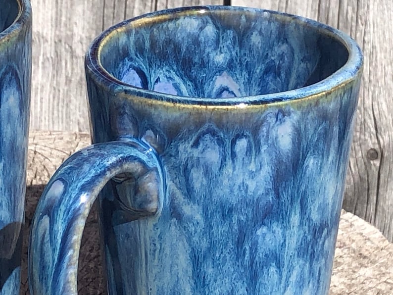 May include: A blue ceramic mug with a marbled glaze. The mug has a wide, rounded handle and a slightly flared rim.