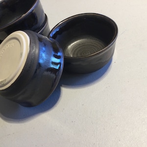 May include: Four black ceramic bowls with a glossy finish. The bowls are stacked on top of each other. The bottom bowl has a white label with the word "Japan" on it.