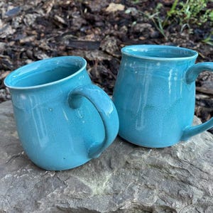 May include: Two handmade ceramic mugs with a glossy turquoise glaze. Each mug features a rounded body, a wide rim, and a curved handle on the side. The finish shows subtle variations in colour intensity, creating a smooth, reflective surface texture.