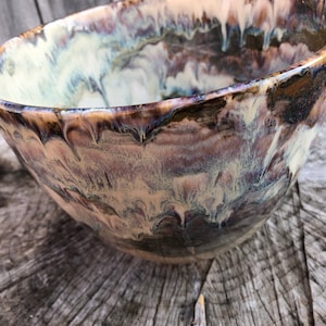 Peut inclure: Un grand bol en céramique avec un glaçage marbré blanc, brun et violet. Le bol a un bord large et arrondi et une base profonde et arrondie.