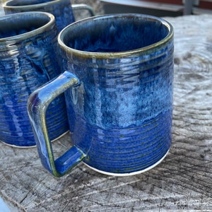 May include: Three blue ceramic mugs with a textured glaze. The mugs have a light blue and dark blue colour variation. The mugs are sitting on a wooden surface.