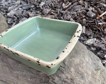 Pode incluir: Uma assadeira quadrada de cerâmica verde claro com uma borda salpicada de branco sujo. A assadeira está vazia e fica sobre uma superfície de pedra cinza, com um fundo de lascas de madeira.