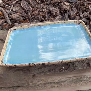 May include: Rectangular ceramic serving tray. The tray has a light blue interior and a textured, tan rim with dark brown accents. The glossy finish makes it suitable for serving food or as a decorative piece.