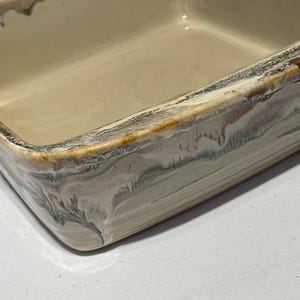 May include: A rectangular ceramic baking dish. It has a cream-coloured interior and a textured, multi-toned rim. The rim features shades of brown, grey, and white, creating a rustic aesthetic. The dish is empty and ready for use.