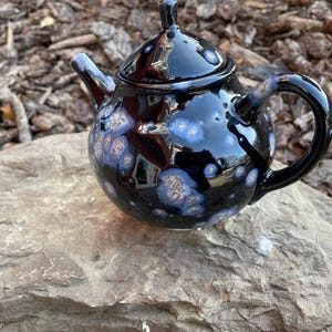 May include: A black teapot with a speckled glaze in shades of blue and white. The teapot has a round body and a small spout. The handle is curved and attached to the side of the teapot.