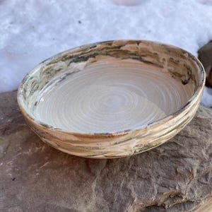 May include: A handmade ceramic bowl with a light beige interior and a textured, swirled design. The rim features brown, green, and beige tones. The bowl is set on a natural stone surface, suggesting a rustic aesthetic.