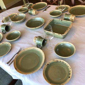 May include: A table setting with green and cream speckled ceramic dinnerware, bowls, and mugs. The table is set with forks and knives.