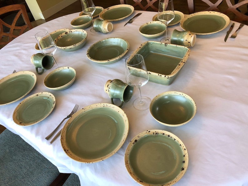 May include: A table setting with green and cream speckled ceramic dinnerware, bowls, and mugs. The table is set with forks and knives.
