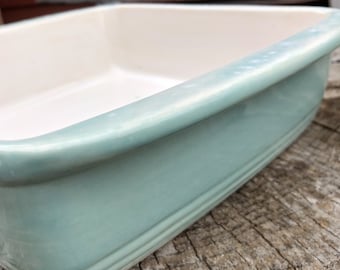 Pode incluir: Uma assadeira retangular com exterior verde-azulado claro e interior branco. A assadeira tem um acabamento liso e brilhante e uma borda ligeiramente curva. O exterior apresenta sulcos horizontais sutis perto da base.