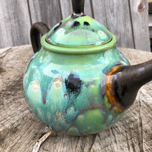 May include: A green and brown ceramic teapot with a lid. The teapot has a speckled glaze with black and gold accents.