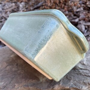 May include: A green ceramic loaf pan with a light green glaze. The pan has a slightly textured surface and a rounded edge.
