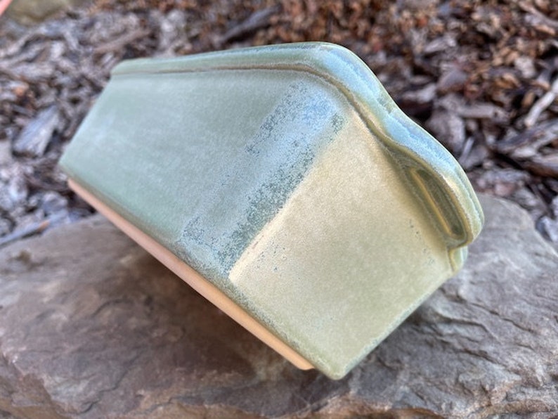 May include: A green ceramic loaf pan with a light green glaze. The pan has a slightly textured surface and a rounded edge.