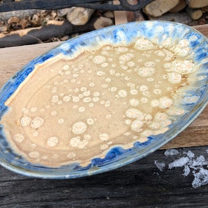 May include: An oval ceramic serving platter in light blue and beige with a speckled glaze. The platter has a raised rim and a slightly irregular shape.