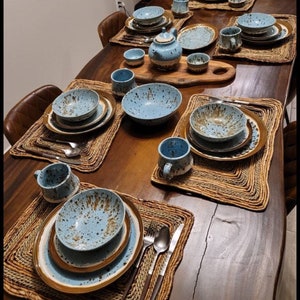 May include: A wooden table set with blue and brown speckled ceramic dinnerware, bowls, mugs, and a teapot. The table is set with woven placemats and includes silverware.