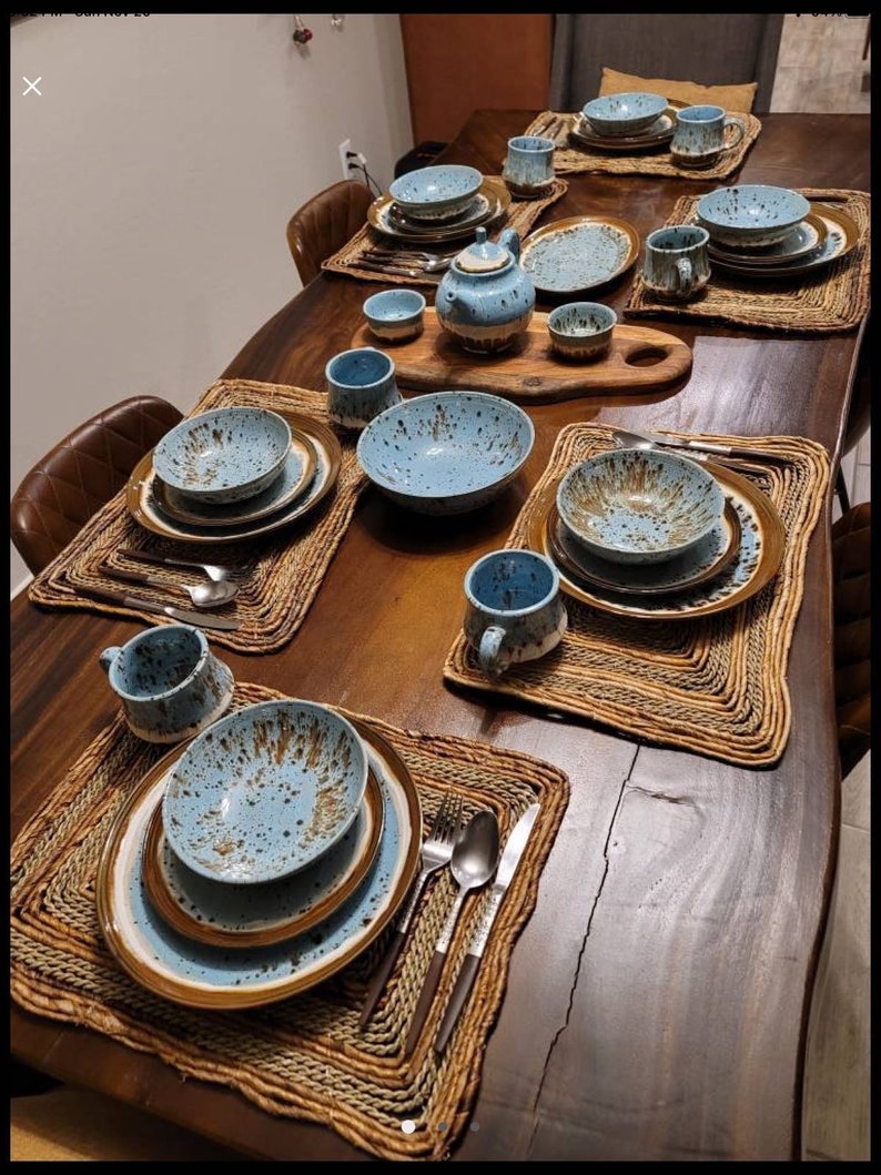 May include: A wooden table set with blue and brown speckled ceramic dinnerware, bowls, mugs, and a teapot. The table is set with woven placemats and includes silverware.