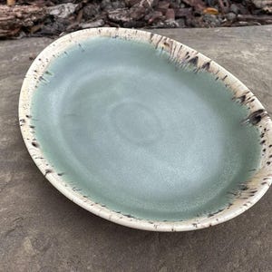 May include: An oval ceramic dish featuring a smooth, sage green centre glaze. The wide, raised rim is finished in a cream-coloured glaze with dark brown, speckled, and streaked accents that radiate inward towards the basin of the plate.