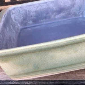 May include: A rectangular baking dish with a light green exterior and a blue interior. The dish has rounded corners and a slightly raised rim. The dish is sitting on a wooden surface.
