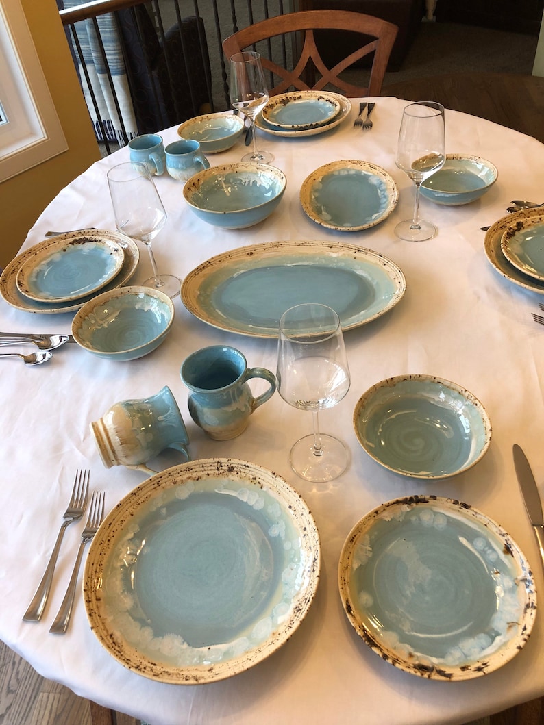 May include: A table setting with blue and white ceramic plates, bowls, and mugs. The plates have a rustic, speckled glaze and a cream-colored rim. There are also wine glasses and silverware on the table.