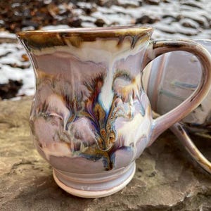 May include: Two ceramic mugs with a unique, marbled glaze in shades of purple, brown, and white. The mugs have a rounded body and a handle.