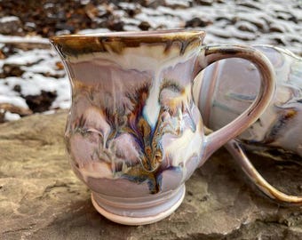 May include: Two ceramic mugs with a unique, marbled glaze in shades of purple, brown, and white. The mugs have a rounded body and a handle.