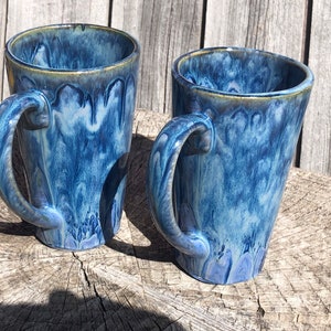 May include: Two blue ceramic mugs with a marbled glaze. The mugs have a tall, tapered shape and a handle on the side.