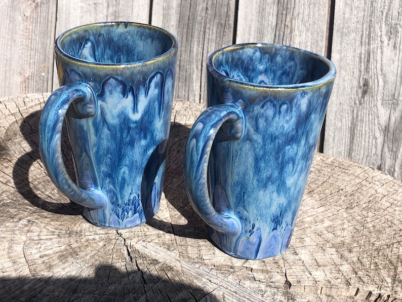 May include: Two blue ceramic mugs with a marbled glaze. The mugs have a tall, tapered shape and a handle on the side.