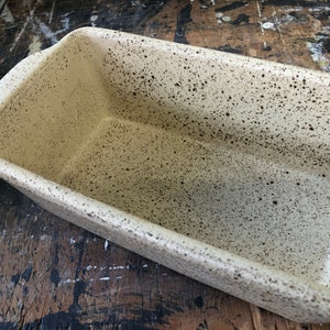 Peut inclure: Un moule à pain en céramique blanc moucheté avec un glaçage légèrement blanc cassé. Le moule a une forme rectangulaire avec des bords arrondis et une poignée sur un côté.
