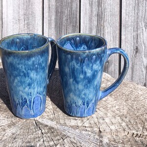 May include: Two blue ceramic mugs with a white glaze and a drip effect. The mugs are sitting on a wooden surface.
