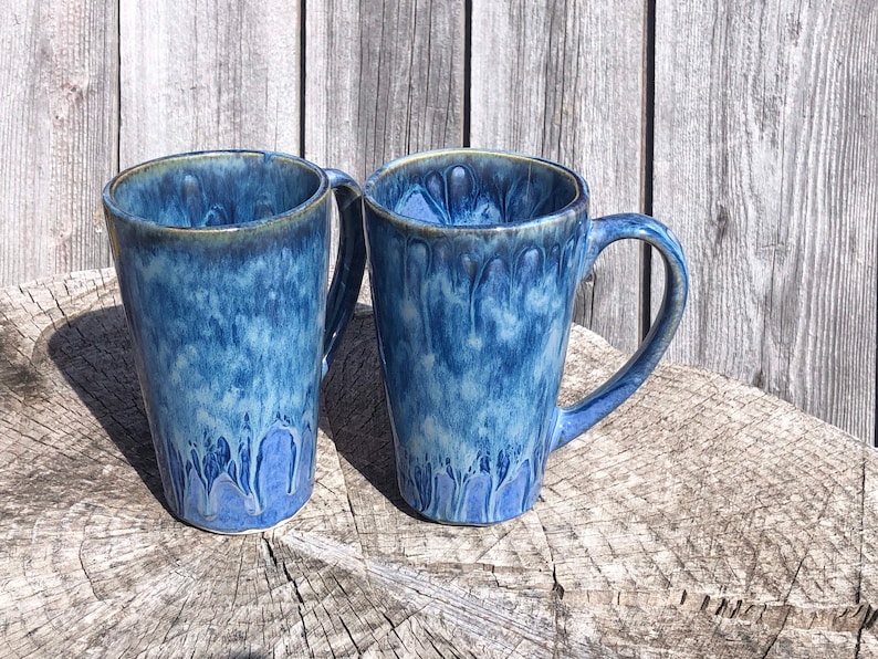 May include: Two blue ceramic mugs with a white glaze and a drip effect. The mugs are sitting on a wooden surface.