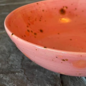 Peut inclure: Un grand bol en céramique rose avec un glaçage moucheté. Le bol a une forme légèrement ovale et est parfait pour servir des salades, des pâtes ou des fruits.