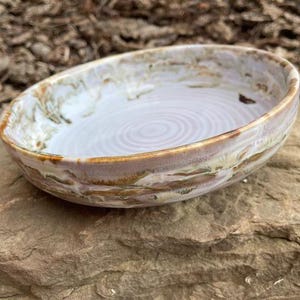 May include: Oval ceramic bowl with a white interior and a swirling pattern. The rim features a blend of brown, beige, and green hues. The bowl is resting on a rough, brown stone surface.