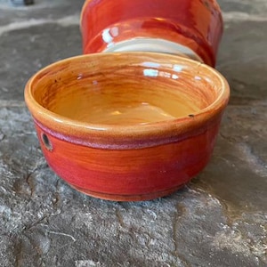 May include: A small, red ceramic bowl with a glazed finish. The bowl has a rounded shape and a slightly flared rim. The inside of the bowl is a lighter shade of orange.